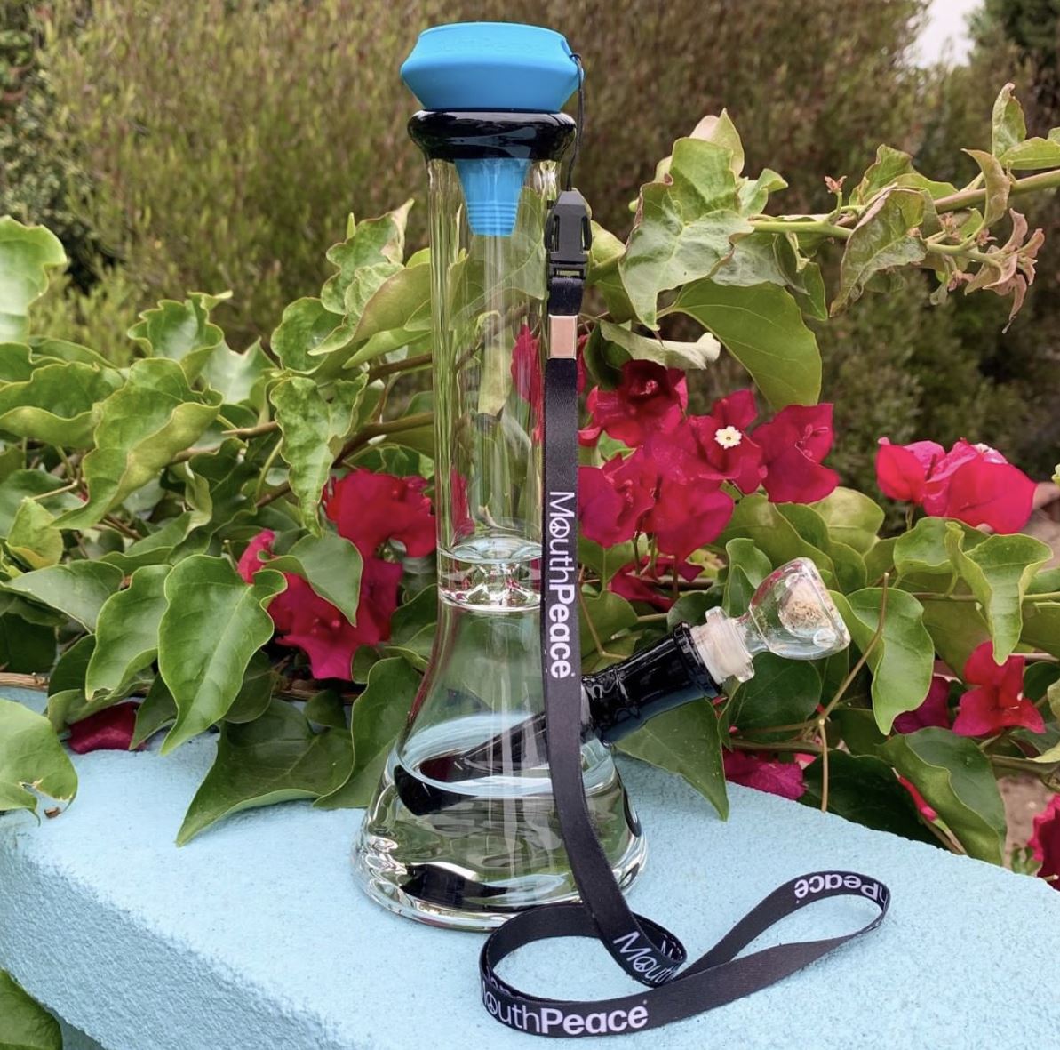 Clear glass bong with a blue silicone MouthPeace filter attached, resting on a ledge against a backdrop of flowers.