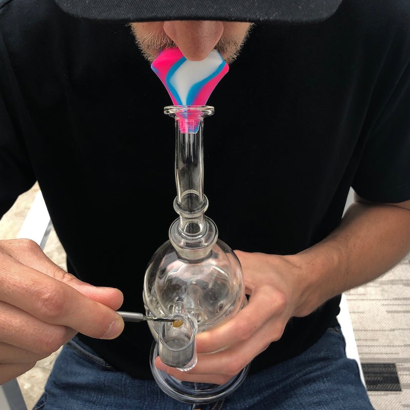 Person using a clear glass bong with a pink and blue silicone mouthpiece to take a hit.