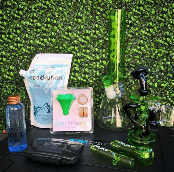 Green glass bongs, a MouthPeace filter, and cleaning supplies arranged on a black surface against a green hedge backdrop.