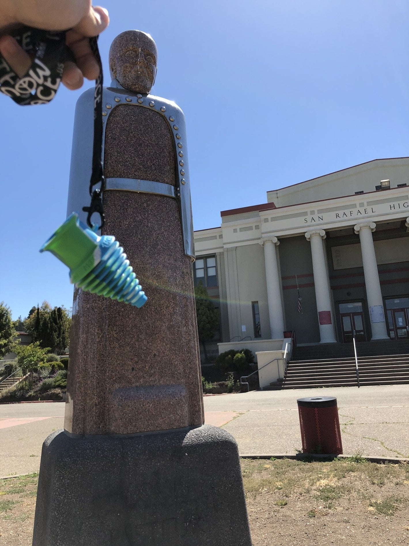 A hand holding a green and blue silicone pipe in front of the historic San Rafael High School statue.